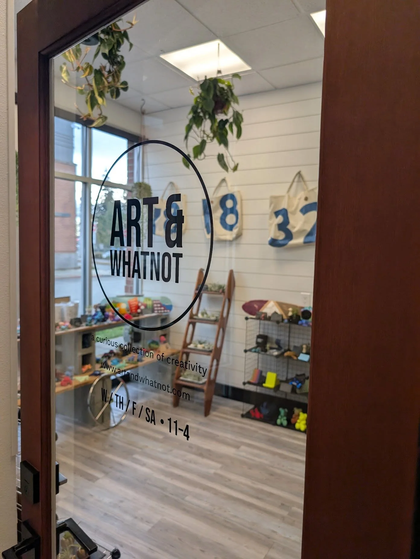 The Art & WhatNot front door with the logo decal, the tagline a curious collection of creativity, the web address, and shop hours, looking into the shop with sail bags, plants, and maker goods on display